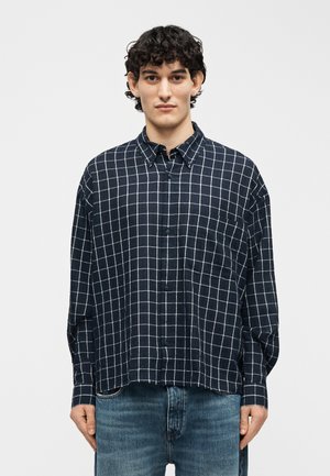 Jeune homme aux cheveux bouclés portant une chemise à carreaux bleu marine à boutons et un jean bleu, debout devant un fond blanc uni.