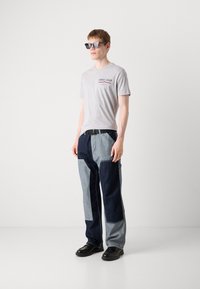 Szürke póló, rajta a "GANT 1949" felirattal, párosítva kéttónusú farmernadrággal, amely sötét és világos árnyalatokban játszik, és fekete slip-on cipőkkel.