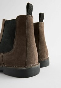 Bottines en suède marron avec panneaux élastiques sur les côtés et semelles en caoutchouc noir. Présente un tissu texturé et des détails de couture sur les bords.