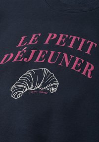 Tissu foncé avec texte rose "LE PETIT DÉJEUNER" au-dessus d'un dessin blanc en lignes d'un croissant avec un petit texte en italique en dessous.
