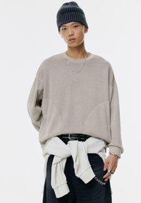Felpa beige oversize con dettagli delle cuciture, indossata sopra una camicia bianca annodata. Pantaloni neri, accessori in catena argentata e un berretto a coste blu navy.