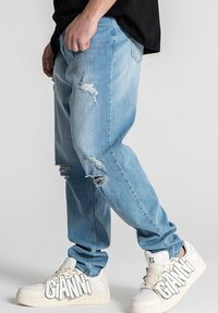 Jeans de color azul claro, rotos y con un ajuste relajado, que presentan detalles desgastados en las rodillas. Llevados con zapatillas blancas que muestran letras en negrita.