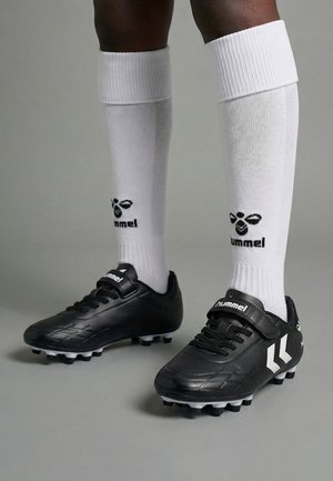 Person iført hvide hummel knæhøje sportssokker og sorte hummel fodboldstøvler med knopper på en grå overflade.