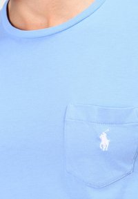 Světle modré bavlněné tričko s kulatým výstřihem a náprsní kapsou s malým bílým logem. Hladká textura a ležérní design.