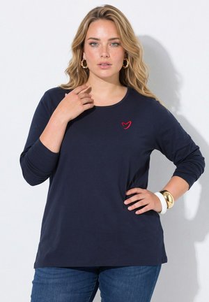 Frau mit langen blonden Haaren, die ein marineblaues Langarmshirt mit kleinem rotem Herzlogo und blaue Jeans trägt, kombiniert mit goldenen Ohrringen und Armbändern.