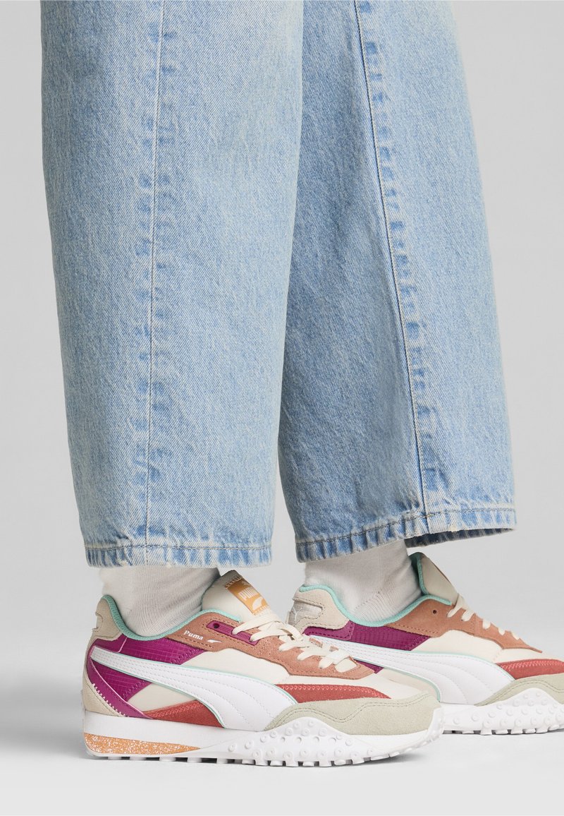 Le sneakers colorate presentano una combinazione di beige, viola e accenti menta con una suola testurizzata. Abbinati a jeans a gamba larga di un blu chiaro.