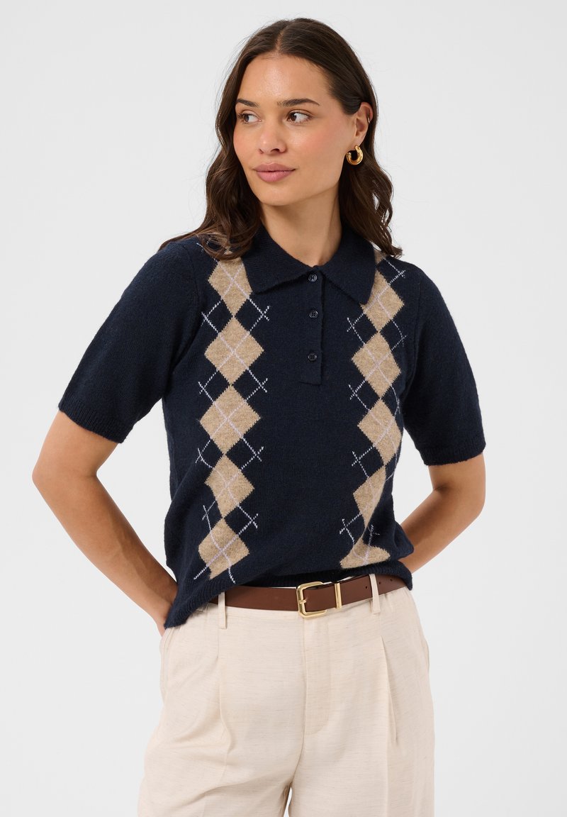 Femme aux cheveux bruns ondulés porte un pull marine à manches courtes à motif argyle, un pantalon beige et des boucles d’oreilles créoles dorées. Elle regarde sur le côté.