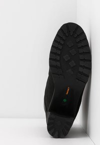 Semelle d'une botte noire avec des motifs de bande de roulement profonds, inclinée contre un angle blanc formé par un mur et un sol.