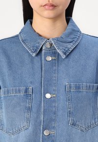Camisa de ganga com um tom azul, gola clássica, fecho de botão, dois bolsos frontais e detalhes desgastados para uma aparência texturizada.