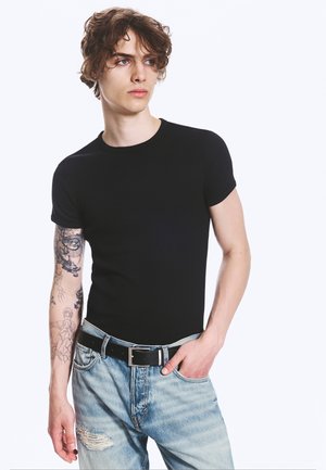 Junge Person mit lockigem Haar trägt schwarzes T-Shirt, helle zerrissene Jeans und einen schwarzen Gürtel, mit detaillierten Tätowierungen am linken Arm, schaut zur Seite.