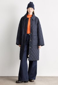 Μακρύ μπουφάν σε ναυτικό μπλε χρώμα με ραμμένο σχέδιο και κούμπωμα με κουμπιά πιέσεως, φορεμένο πάνω από ένα πορτοκαλί ριμπ πλεκτό πουλόβερ και συνδυασμένο με τζιν παντελόνι με φαρδιά πόδια.