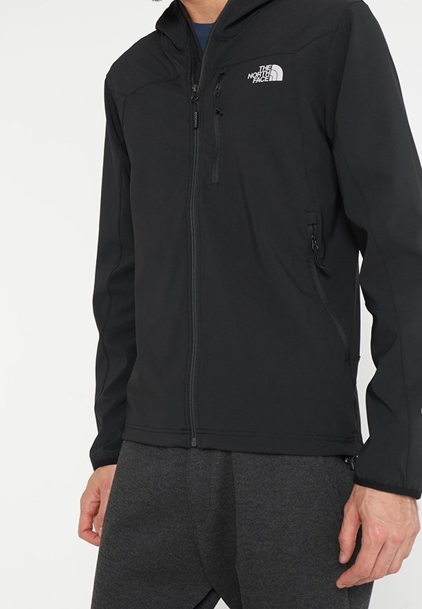 Personne portant une veste softshell noire North Face zippée avec poches zippées sur la poitrine et les côtés, associée à un pantalon gris foncé.