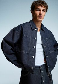 Giacca in denim scuro con vestibilità rilassata, tasche frontali e bottoni argentati. Indossata sopra una canotta chiara, con dettagli cuciti e una leggera lucentezza.