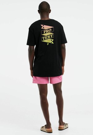 Schwarzes Baumwoll-T-Shirt mit auffälligem, mehrfarbigem "PRO TEST"-Grafik auf dem Rücken, kombiniert mit pinken Shorts. Einfaches Design, lässige Passform.