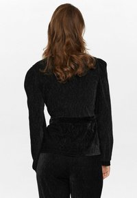 Blazer in velluto a coste nero con tessuto testurizzato, spalle strutturate, vita aderente e maniche lunghe. La vista posteriore mostra linee sinuose e una leggera lucentezza.