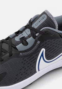 Černá sportovní bota se šedými akcenty, texturovaným svrškem, zakřiveným bílým logem a tmavými tkaničkami. Má polstrovaný límec a výrazný design podrážky.