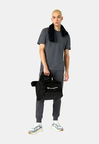 Schwarze Sporttasche mit weißem "Champion"-Logo, gehalten von einer Person in einem grauen Outfit, einschließlich eines kurzärmeligen Shirts und Jogginghosen.
