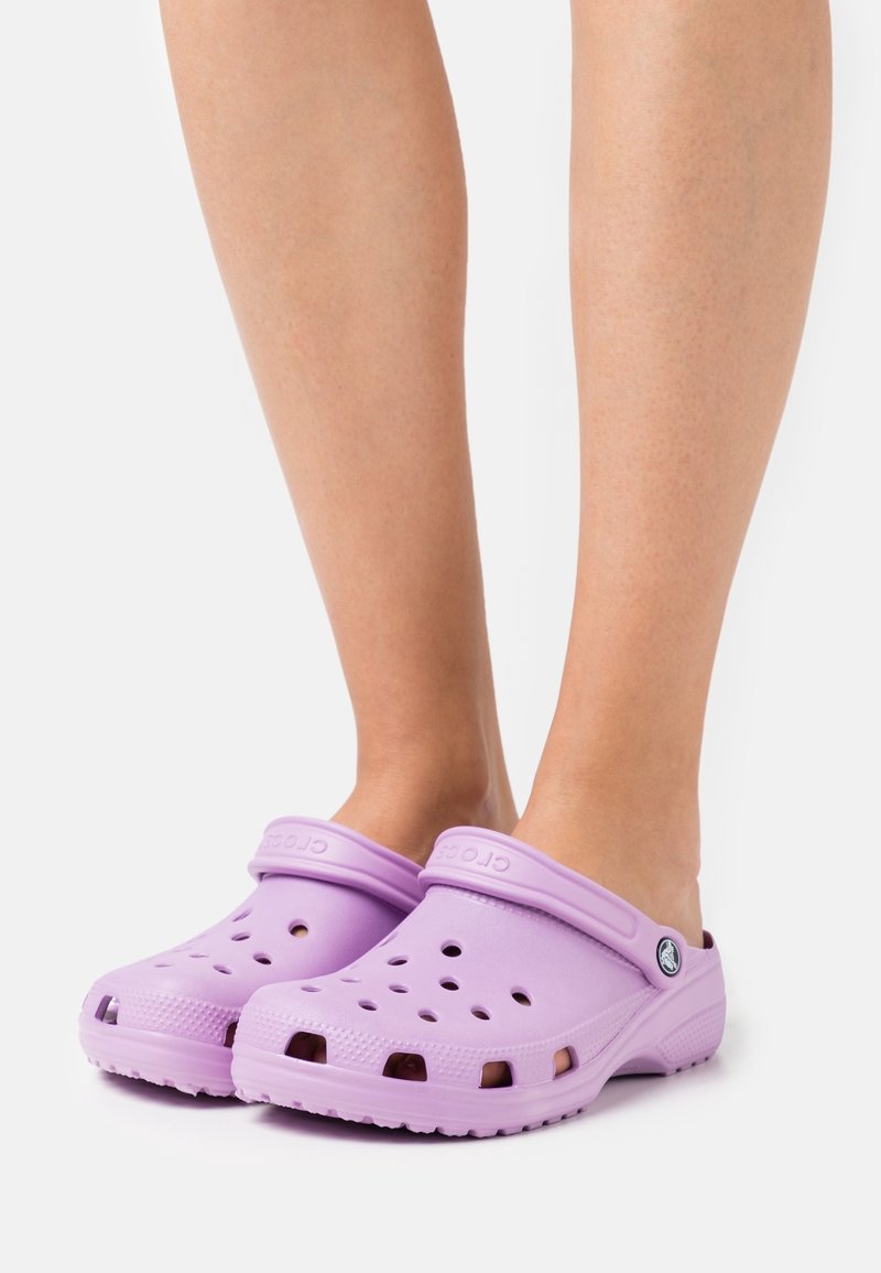 Jambes portant des sabots violet clair à enfiler avec des trous de ventilation et des lanières arrière réglables sur fond blanc.
