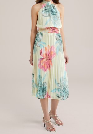 Donna che indossa un abito crema senza maniche con stampa floreale grande rosa e blu e gonna midi plissettata, abbinato a tacchi bianchi con cinturini.