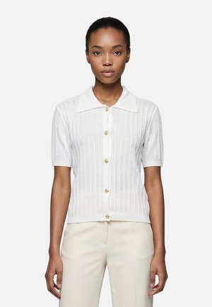Donna che indossa un cardigan a maglia bianco a maniche corte con bottoni dorati e colletto, abbinato a pantaloni beige chiaro, in piedi davanti a uno sfondo semplice.