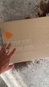 Brauner Karton mit orangefarbenem dreieckigem Logo und weißem Text: "Love me. Wear me." auf einer strukturierten Oberfläche platziert.