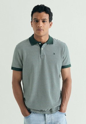 Joven con cabello rizado y oscuro que lleva una camiseta polo de rayas verdes y blancas y vaqueros azul claro, de pie con las manos en los bolsillos frente a un fondo liso.