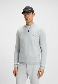 Pullover grigio chiaro a maniche lunghe con mezza cerniera, dettagli texturizzati e un logo nero. Abbinato a pantaloni grigi chiari e un cappellino nero.