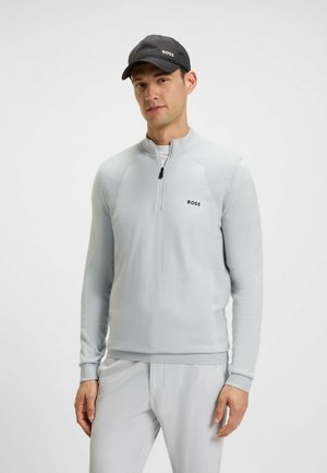 Light gray long-sleeve pullover with half-zip, textured accents, and a black logo. Paired with light gray pants and a black cap.