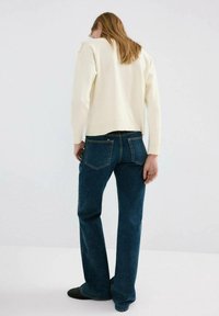 Pulover tricotat de culoare crem, croială lejeră, asortat cu blugi din denim albastru închis, cu design clasic cu cinci buzunare. Încălțăminte cu talpă plată vizibilă.