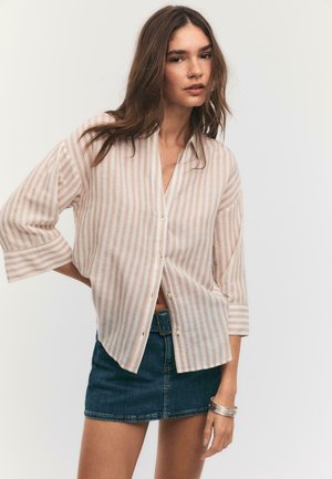 Mladá žena s dlhými hnedými vlasmi, oblečená v béžovo-bielej prúžkovanej košeli na gombíky a tmavomodrej denimovej mini sukni.