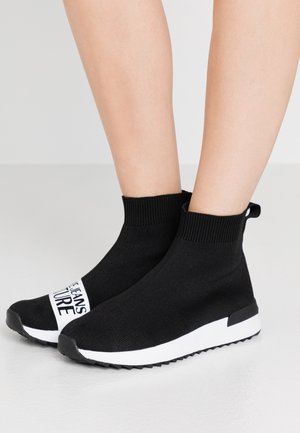 Czarne sneakersy typu skarpetkowego z białymi podeszwami i detalem tekstowym noszone na nagich nogach na jednolitym jasnym tle.