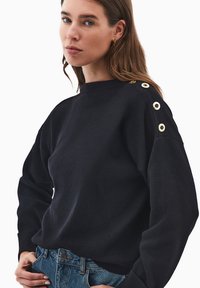 Schwarzer Pullover mit Rundhalsausschnitt, ausgestattet mit goldenen Knopf-Details an der linken Schulter. Aus weichem Material gefertigt, mit einem lockeren Schnitt und geripptem Saum.