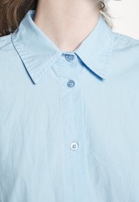 Camicia azzurra chiaro con colletto e chiusura a bottoni frontale, realizzata in tessuto morbido, con una sottile texture e dettagli di cucitura appena visibili.