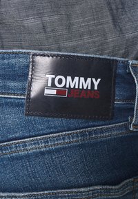 Niebieskie dżinsy z denimu z ciemną skórzaną naszywką z napisem "TOMMY JEANS" w białym i czerwonym kolorze. Widoczne szwy i faktura tkaniny.