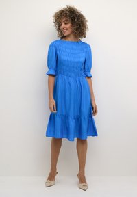 Rochie albastră cu elastic, cu mâneci scurte și pufoase, tiv ondulat și o textură netedă și lucioasă. Rochia se termină puțin sub genunchi.
