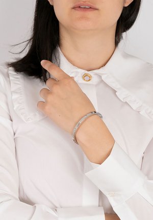 Bracelet en argent avec des accents circulaires, porté sur une main. Le porteur porte une blouse blanche à volants avec un bouton doré rond au col.