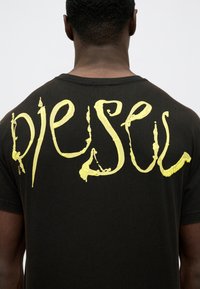 Vyras dėvi juodą marškinėlį su didelėmis geltonomis tekstūruotomis raidėmis, rašančiomis "PIEUSA" ant nugaros, priešais vientisą foną.