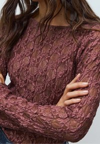 Gros plan sur une femme portant un haut à manches longues en dentelle florale mauve avec un col rond, les bras croisés sur sa poitrine.