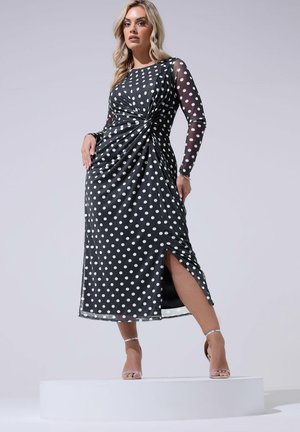 Frau steht auf weißer Plattform und trägt ein schwarzes Midikleid mit weißen Punkten, durchsichtigen langen Ärmeln, Seitenschlitz und silbernen High-Heel-Sandalen.