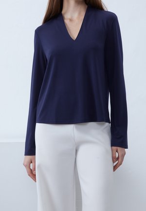 Woman wearing a navy blue long-sleeve V-neck top and white pants, standing against a plain light background.