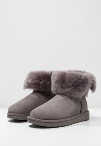 Stivaletti alla caviglia in suede grigio con un morbido risvolto in pelliccia sintetica grigia. Superficie texturizzata e suola piatta in gomma con un leggero motivo di battistrada.