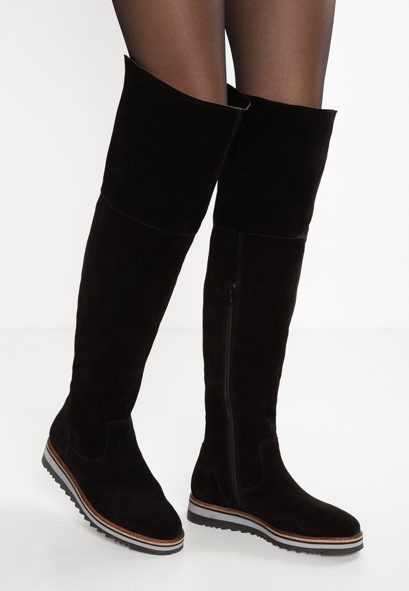 Bottes hautes en daim noir jusqu'au genou avec fermeture éclair latérale et semelles compensées portées sur des collants transparents foncés, sur fond blanc.