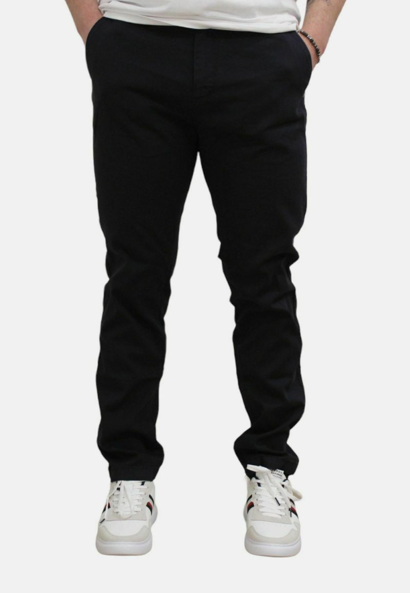 Zwarte slim-fit broek met een gladde textuur, voorzien van zijzakken en een eenvoudig ontwerp, gecombineerd met zwart-witte sneakers.