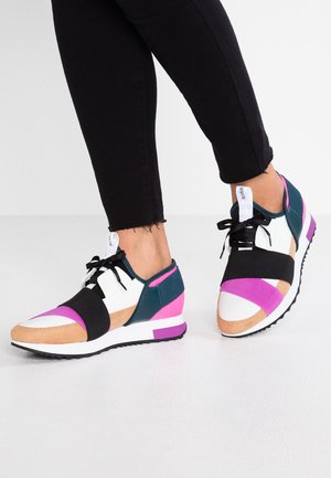 Osoba ubrana w czarne spodnie i kolorowe sneakersy z białymi, różowymi, beżowymi, czarnymi i turkusowymi akcentami, stojąca na jasnej podłodze przy białej ścianie.