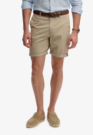 Mann trägt beige hochgekrempelte Shorts, braunen geflochtenen Gürtel, hellblaue hochgekrempelte Hemd, beige Slipper und eine Armbanduhr mit beigem Armband.