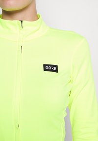 Neon keltainen pitkähihaine paita, jossa on korkea kaulus ja etuvetoketju. Rintataskussa on musta GORE-logo. Suora, vartaloa myötäilevä muotoilu.