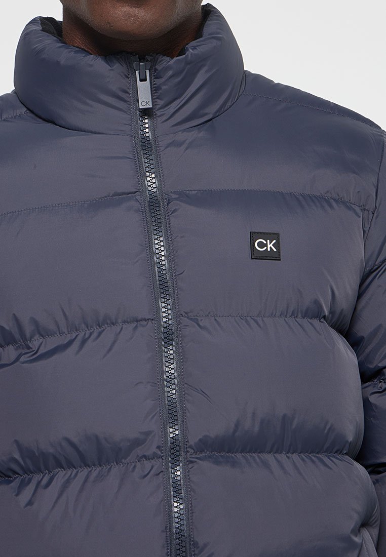 Homme portant une veste matelassée bleu marine avec un col montant et un patch logo CK sur la poitrine, zippée jusqu'au cou.