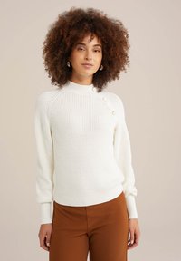 Baltas megztinis su aukšta apykakle, raukšlėtais rankogaliais ir prigludusia apatine dalimi. Kairėje peties pusėje yra dekoro mygtukai. Lygus paviršius.