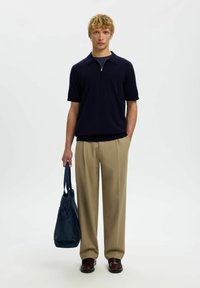 Jeune homme portant une chemise à manches courtes bleu marine avec col zippé, pantalon beige, tenant un sac fourre-tout bleu marine, debout devant un fond blanc uni.