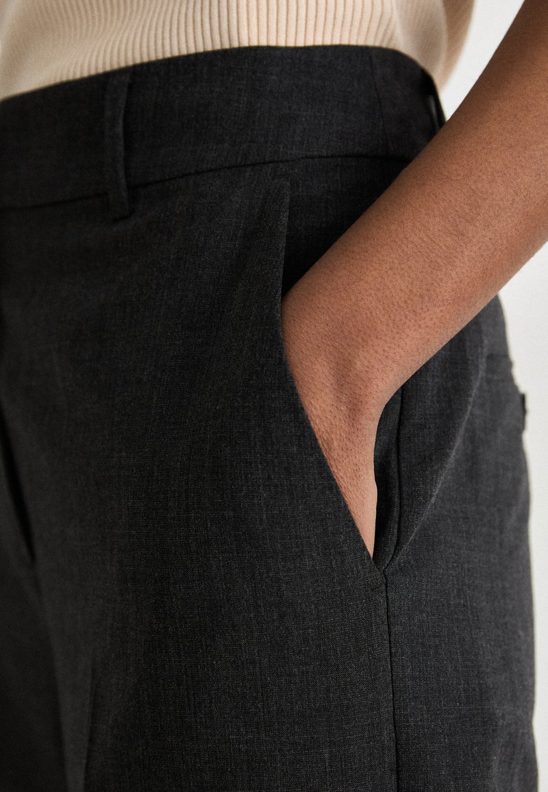 Dunkelgraue Hose aus strukturiertem Stoff, mit Seitentaschen und tailliertem Schnitt, kombiniert mit einem hellen, gerippten Oberteil.